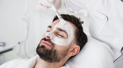 Cosmetologist using brush while putting white clay mask on male face GoldenOrchard